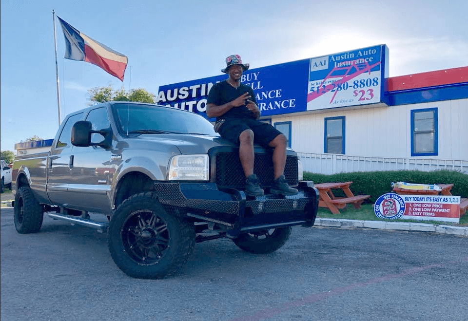 Happy customer sitting on the hood of a large pickup in front of Austin Auto Traders.